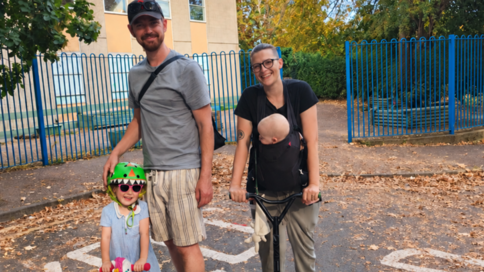 Hebe and her family. She is wearing her little one in a carrier, standing on a scooter and her eldest is on a scooter while Alan stands on a skateboard.