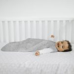 Smiling baby laid in a white cot wearing a grey sleeping bag - choosing a mattress and bedding for a baby